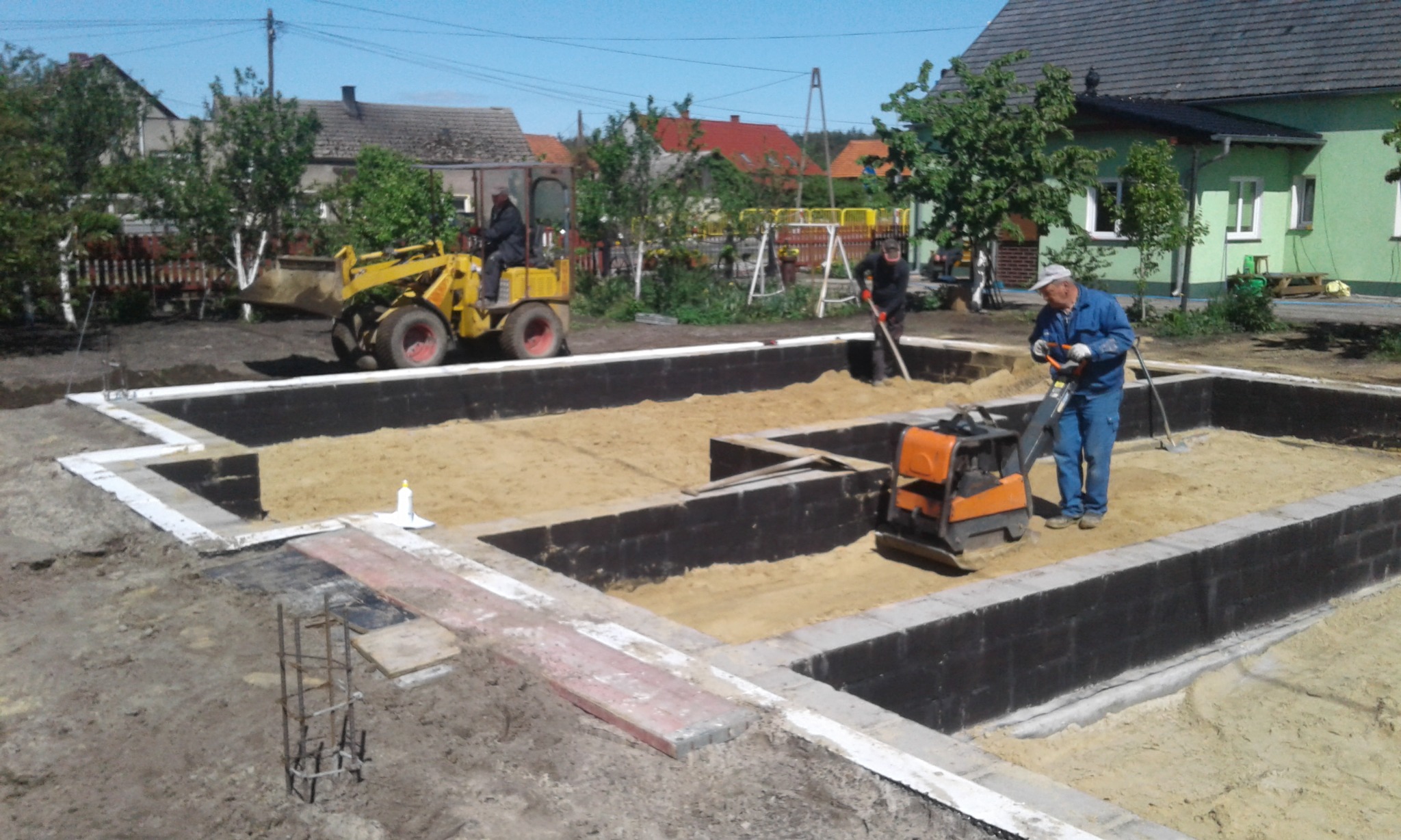 Budowa fundamentów: pracownicy zagęszczają piasek w wykopie pod fundamenty domu jednorodzinnego, widoczny żółty mini-ładowarka i ubijarka wibracyjna.