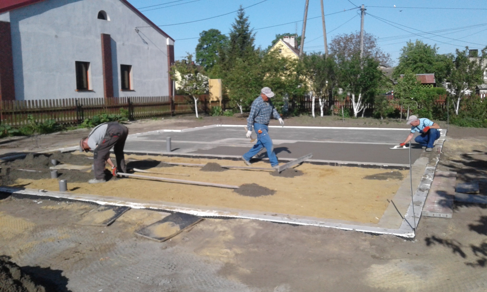 Trzech robotników na budowie w trakcie wylewania fundamentów pod budynek mieszkalny, jeden wyrównuje ziemię, drugi rozprowadza beton, a trzeci wygładza powierzchnię, w tle widoczny budynek...