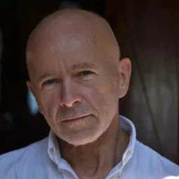 Portret dojrzałego, łysiejącego mężczyzny w białej koszuli, z delikatnym uśmiechem, na tle ciemnego, drewnianego elementu architektonicznego i rozmytej sylwetki.