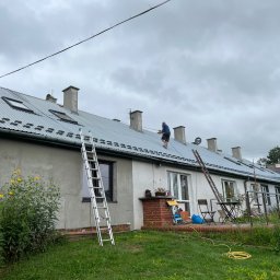 Zając Dachy S.C. J.Zając - Pracownik na dachu szeregowca z blachy trapezowej, zabezpieczony liną, z widocznymi kominami i oknami dachowymi; dwie drabiny opierają się o budynek, a na trawniku widoczne kwiaty i meble ogrodowe.