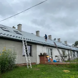 Pracownik na dachu szeregowca z blachy trapezowej, zabezpieczony liną, z widocznymi kominami i oknami dachowymi; dwie drabiny opierają się o budynek, a na trawniku widoczne kwiaty i meble ogrodowe.