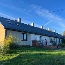 Zając Dachy S.C. J.Zając - Długi, parterowy budynek z szarym dachem z blachy trapezowej i jasną elewacją, widok z trawnika, dwie osoby na tarasie przed budynkiem, błękitne niebo z smugami kondensacyjnymi.