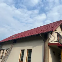 Dom jednorodzinny z nowym, czerwonym dachem z blachodachówki, widoczny z dołu pod kątem, z przystawioną drabiną do rynny i lukarną w dachu. Na ścianie balkon z ozdobną balustradą.