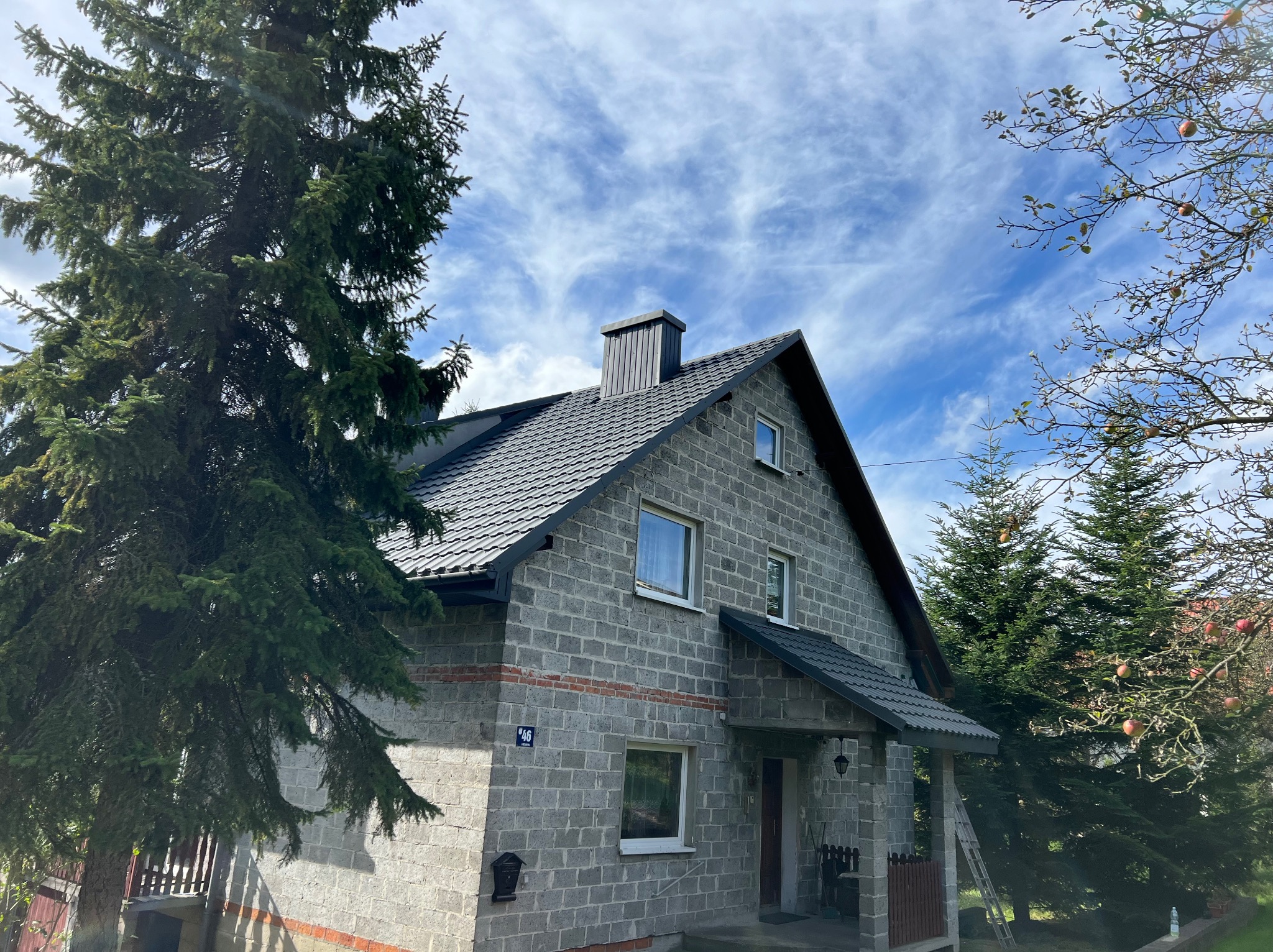 Dom z szarej cegły w Wadowicach z nowym, ciemnym dachem z dachówki, widoczny komin i fragment elewacji, na tle błękitnego nieba z chmurami i zieleni drzew.