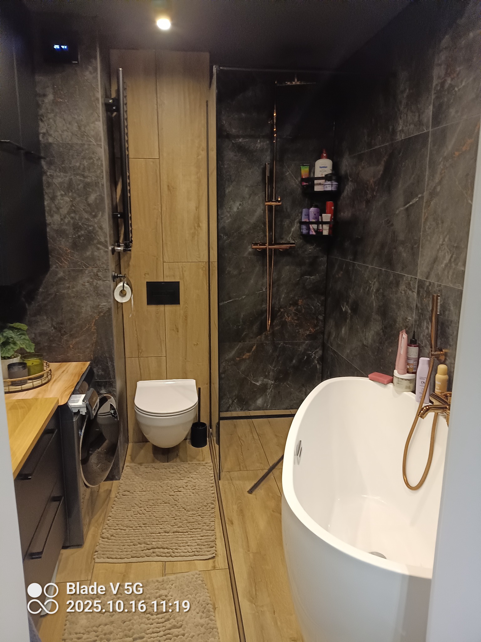 Nowoczesna łazienka z kabiną prysznicową typu walk-in, wolnostojącą wanną i drewnopodobnymi płytkami. Uwagę zwraca armatura w kolorze różowego złota.