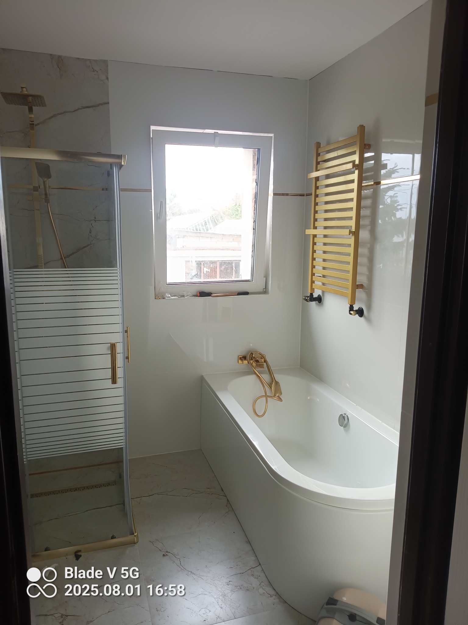 Jasna łazienka po remoncie: biała wanna, kabina prysznicowa z poziomymi paskami, złota armatura i grzejnik drabinkowy. Okno z widokiem na zewnątrz.