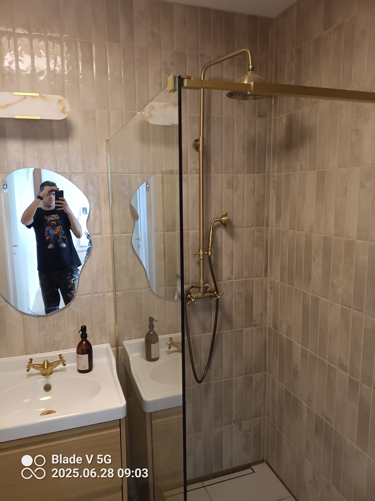 Wykończona łazienka z beżowymi płytkami, złotą armaturą i lustrem o nieregularnym kształcie. Widoczna kabina prysznicowa ze szklaną ścianką i odpływ liniowy.