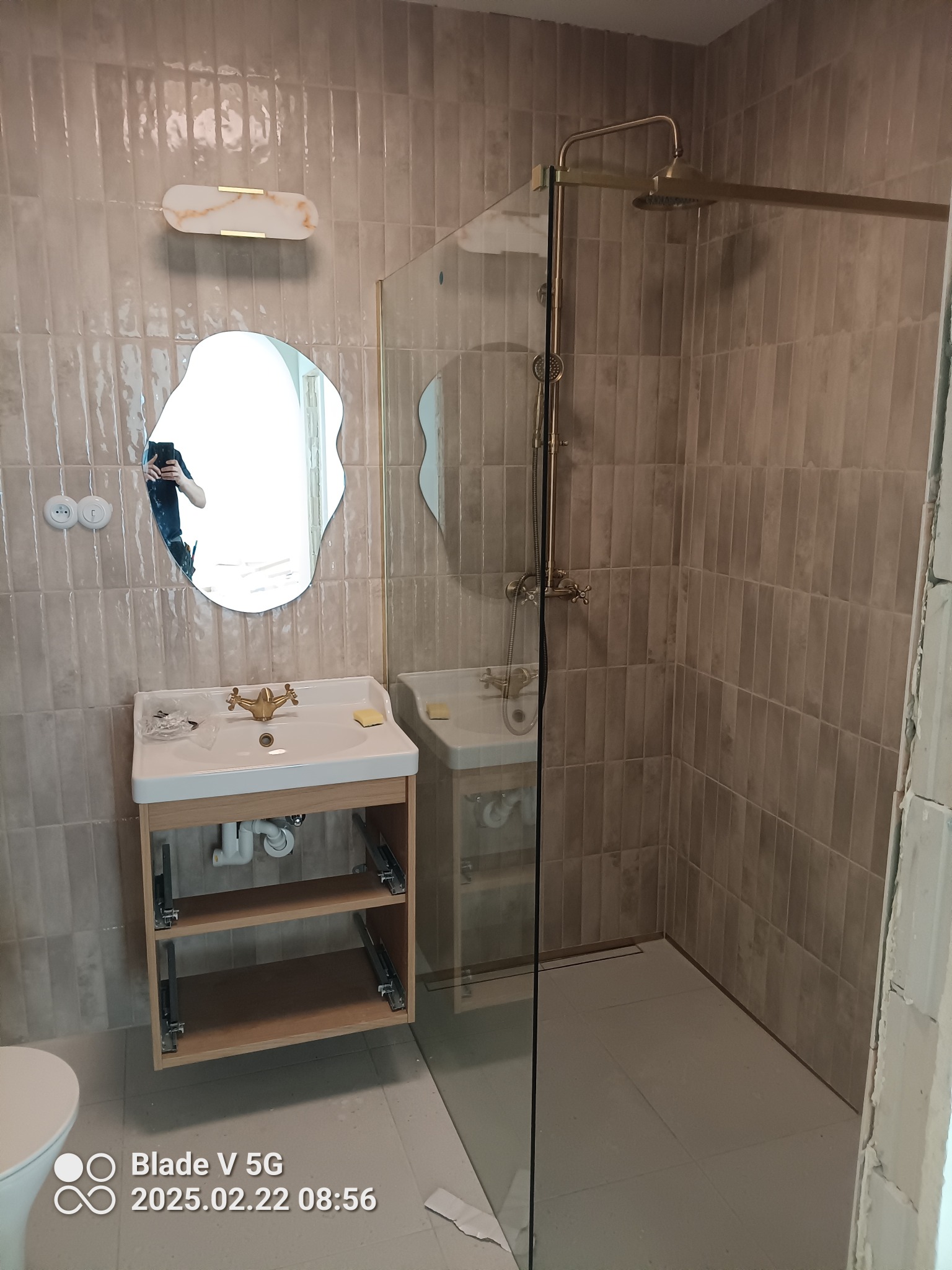 Wykończona łazienka z beżowymi płytkami, kabiną prysznicową ze złotymi elementami i umywalką na drewnianym stelażu. Lustro o nieregularnym kształcie. Widoczne detale wykończeniowe.