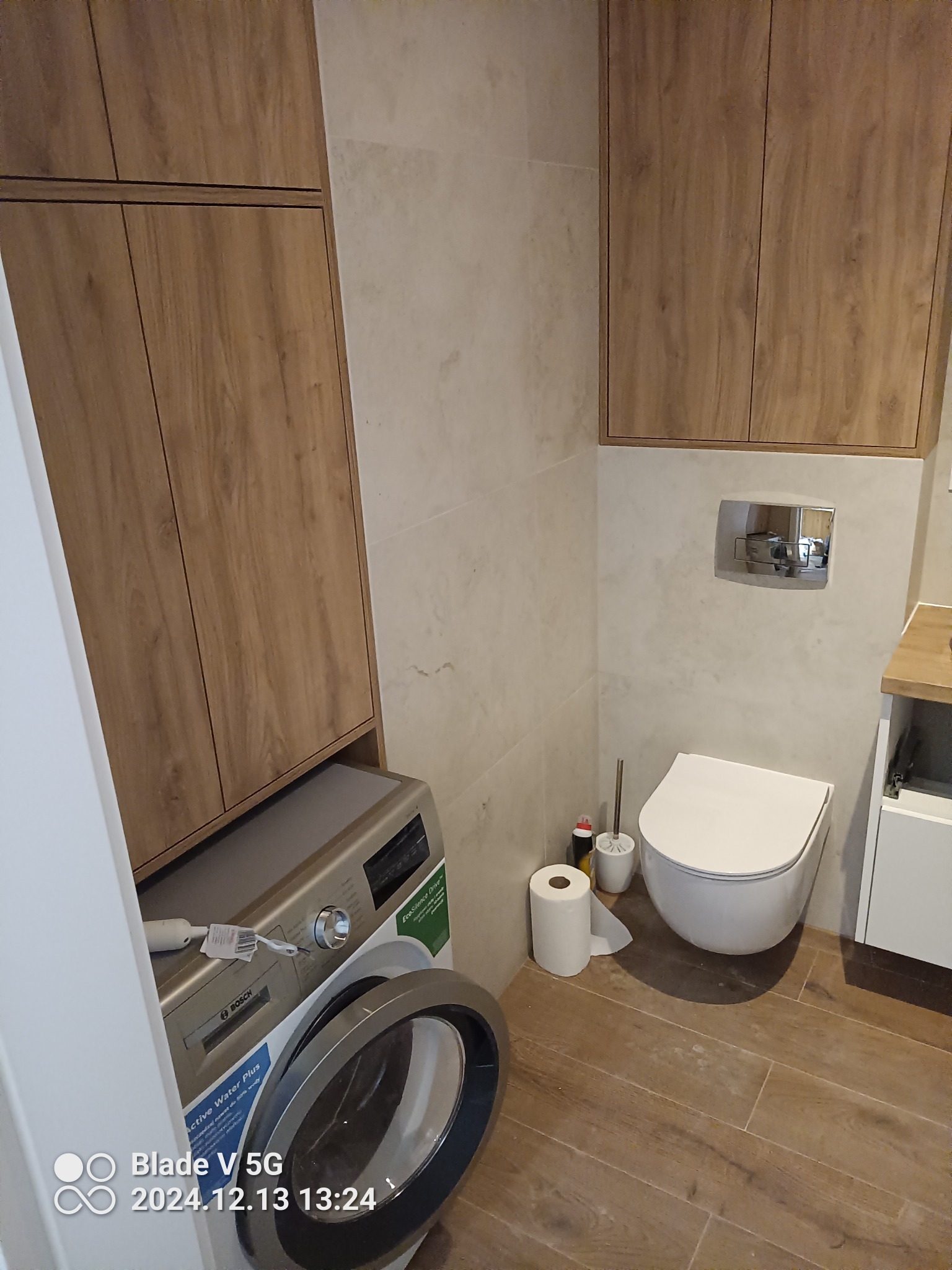 Nowoczesna łazienka z drewnianymi szafkami, beżowymi płytkami i pralką Bosch. Uwagę przykuwa wisząca toaleta i drewnopodobna podłoga.