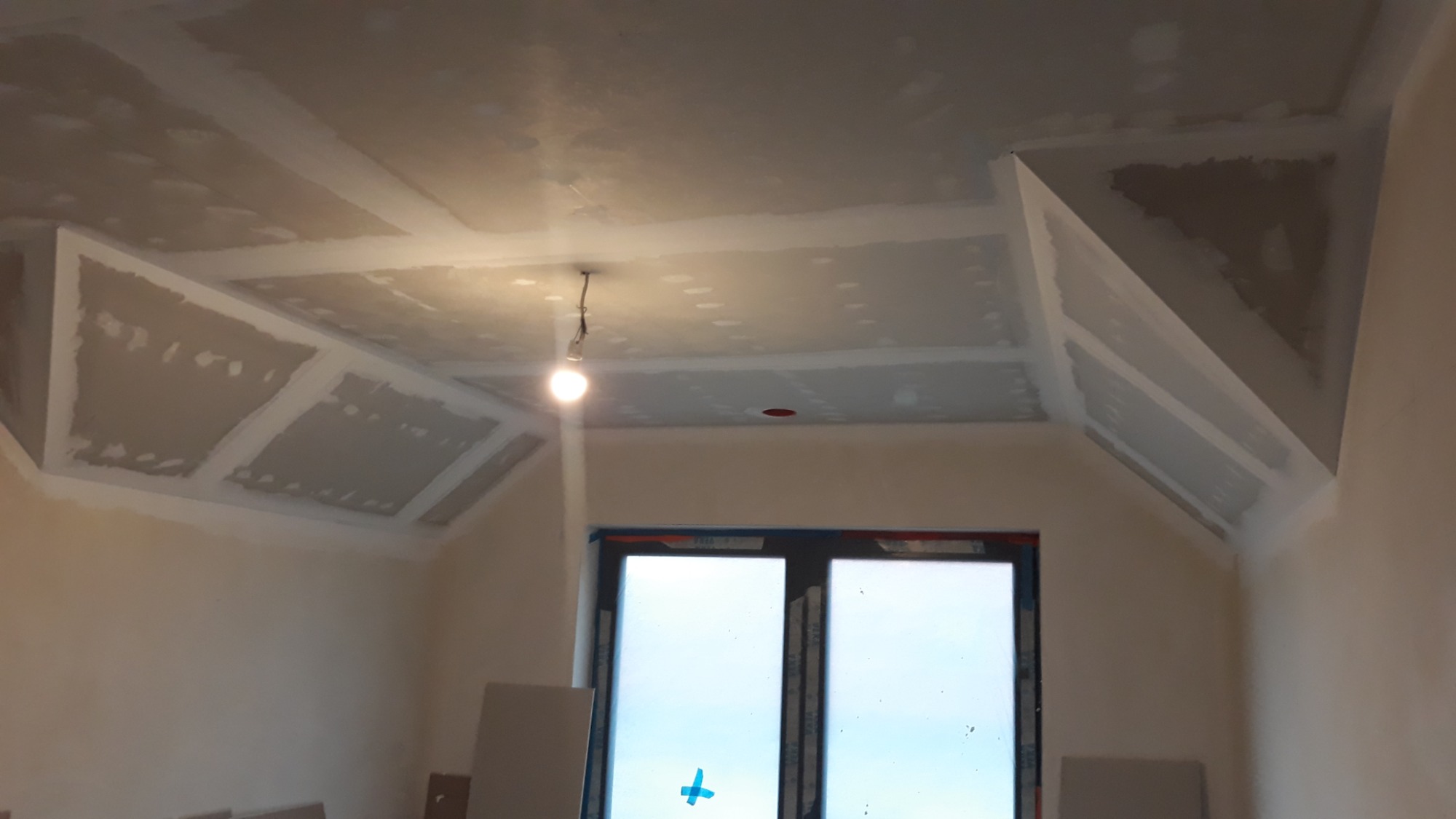 Wykończony sufit z płyt gipsowo-kartonowych z widocznymi łączeniami i narożnikami, oświetlony wiszącą żarówką, okno w tle.