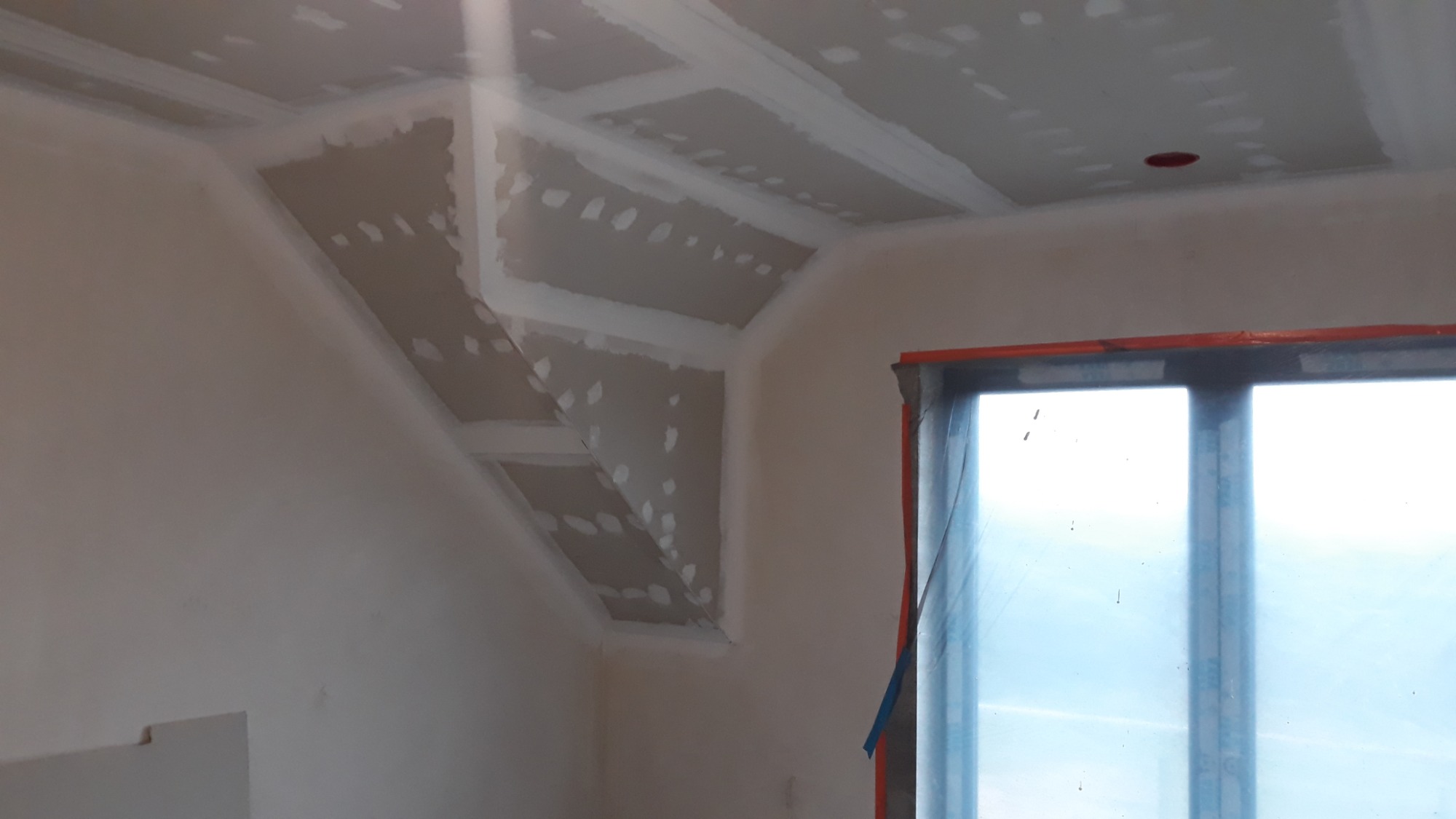 Skośny sufit na poddaszu w trakcie montażu, widoczne płyty zaszpachlowane na łączeniach, okno z folią ochronną, przygotowane do dalszych prac wykończeniowych.