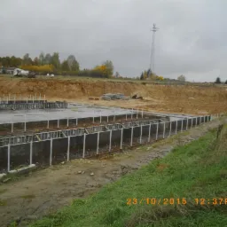 Przygotowanie terenu pod budowę domu: szalunki fundamentowe zbrojone stalą, widoczny wykop ziemny i stosy materiałów budowlanych w tle, pochmurny dzień.