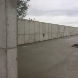 Surowy, betonowy fundament z widocznymi segmentami szalunku i pionowym słupem wspierającym, świeżo wylana posadzka z włazami kanalizacyjnymi, pochmurne niebo w tle.