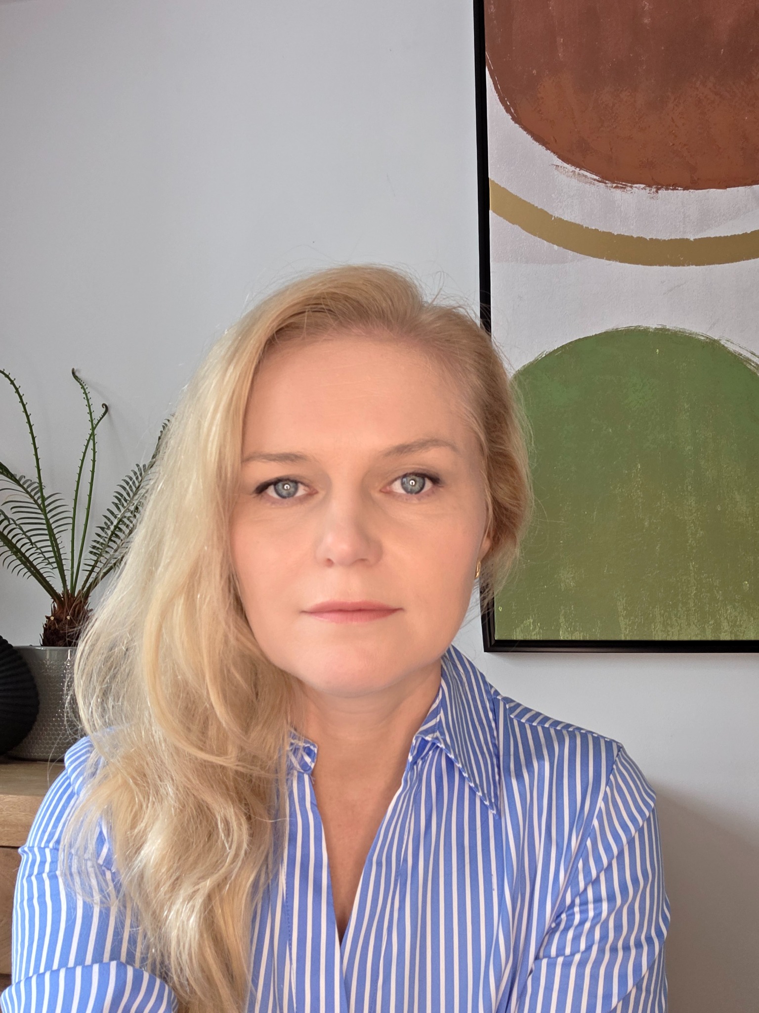 Portret kobiety o blond włosach w niebieskiej koszuli na tle ściany z obrazem i rośliną doniczkową. Ujęcie od przodu, naturalne światło.