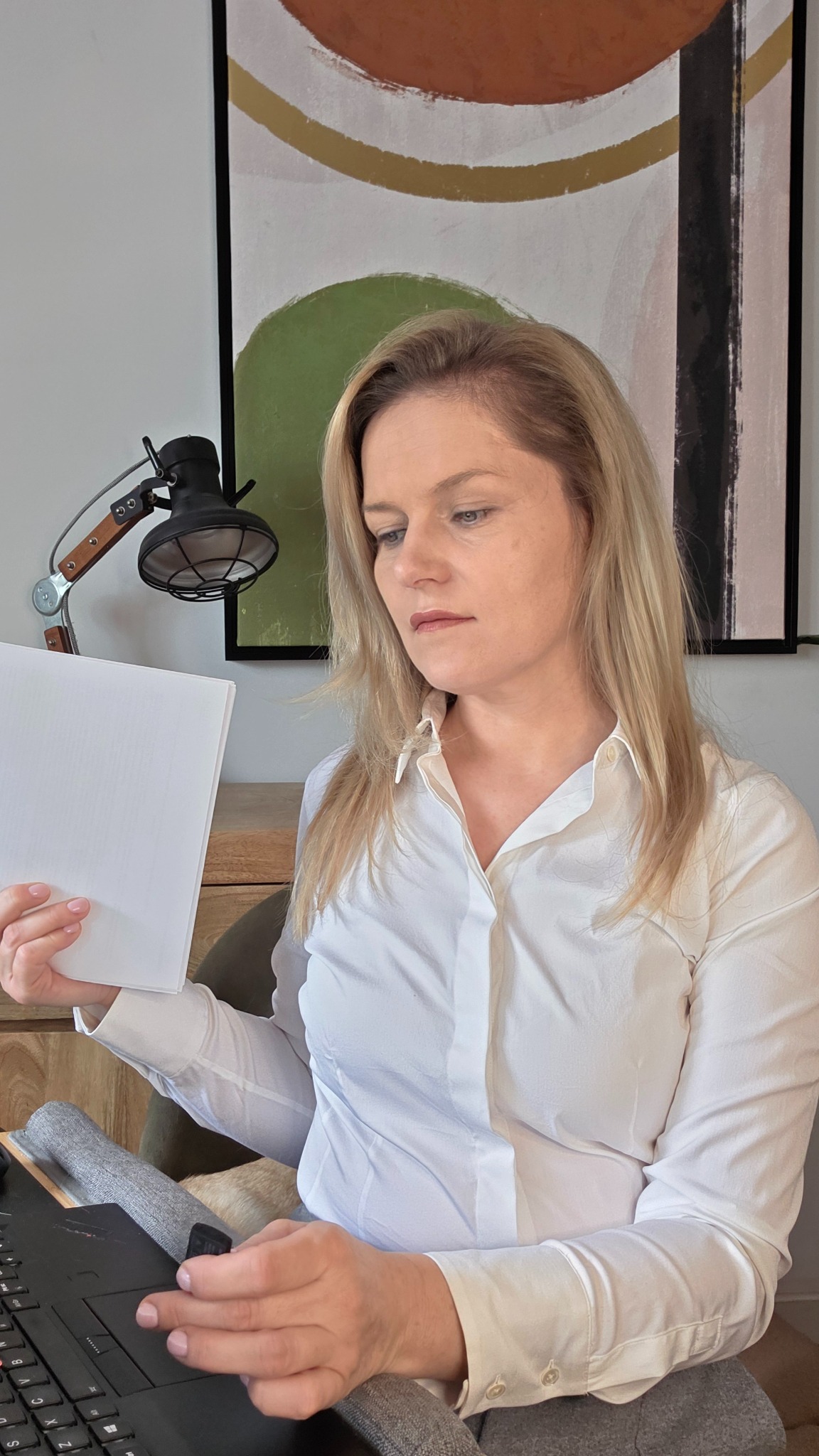 Kobieta w białej koszuli analizuje dokumenty przy biurku z lampką. W tle abstrakcyjny obraz. Laptop widoczny na pierwszym planie.