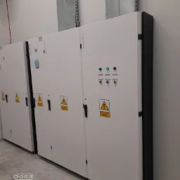 Rząd szaf elektrycznych marki Legrand w jasnej obudowie z czarnymi detalami, z widocznymi oznaczeniami ostrzegawczymi i elementami sterującymi, zamontowanych w pomieszczeniu z szarą podłogą...