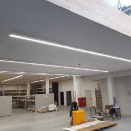 Długie, smukłe lampy LED zamontowane w suficie podwieszanym w hali magazynowej, widoczne regały i materiały budowlane w tle.