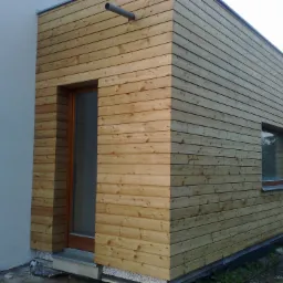 Nowoczesny dom z elewacją z jasnego drewna, widoczny fragment ściany z oknem i drzwiami wejściowymi, rynna odprowadzająca wodę na elewacji.