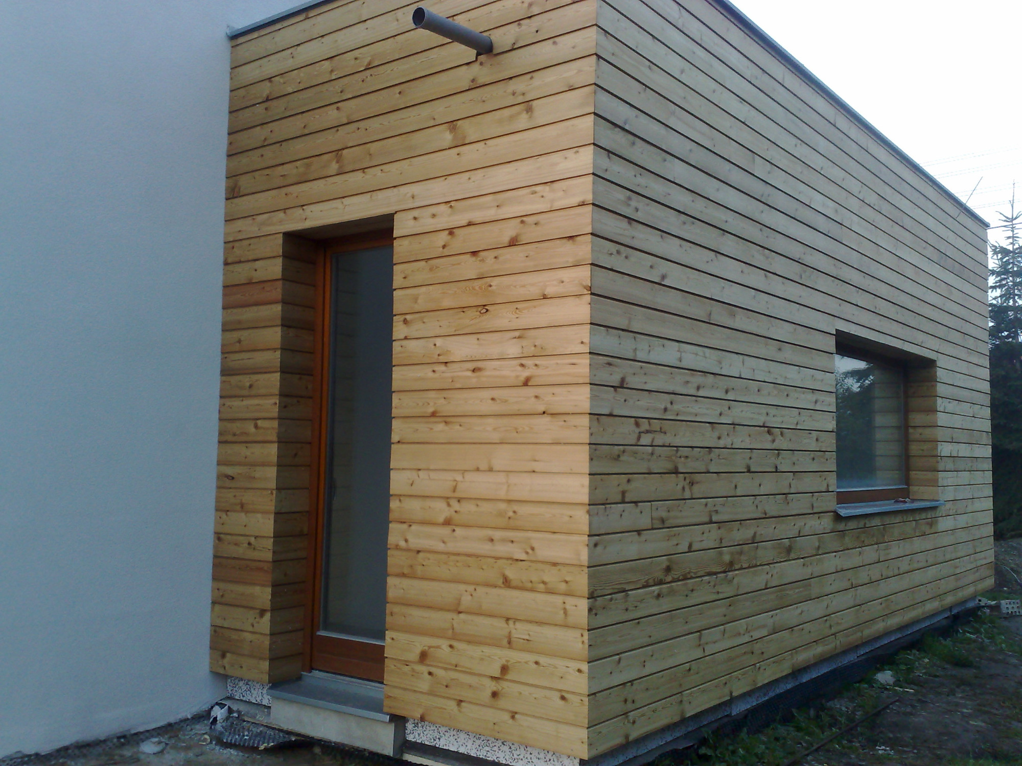 Nowoczesny dom z elewacją z jasnego drewna, widoczny fragment ściany z oknem i drzwiami wejściowymi, rynna odprowadzająca wodę na elewacji.