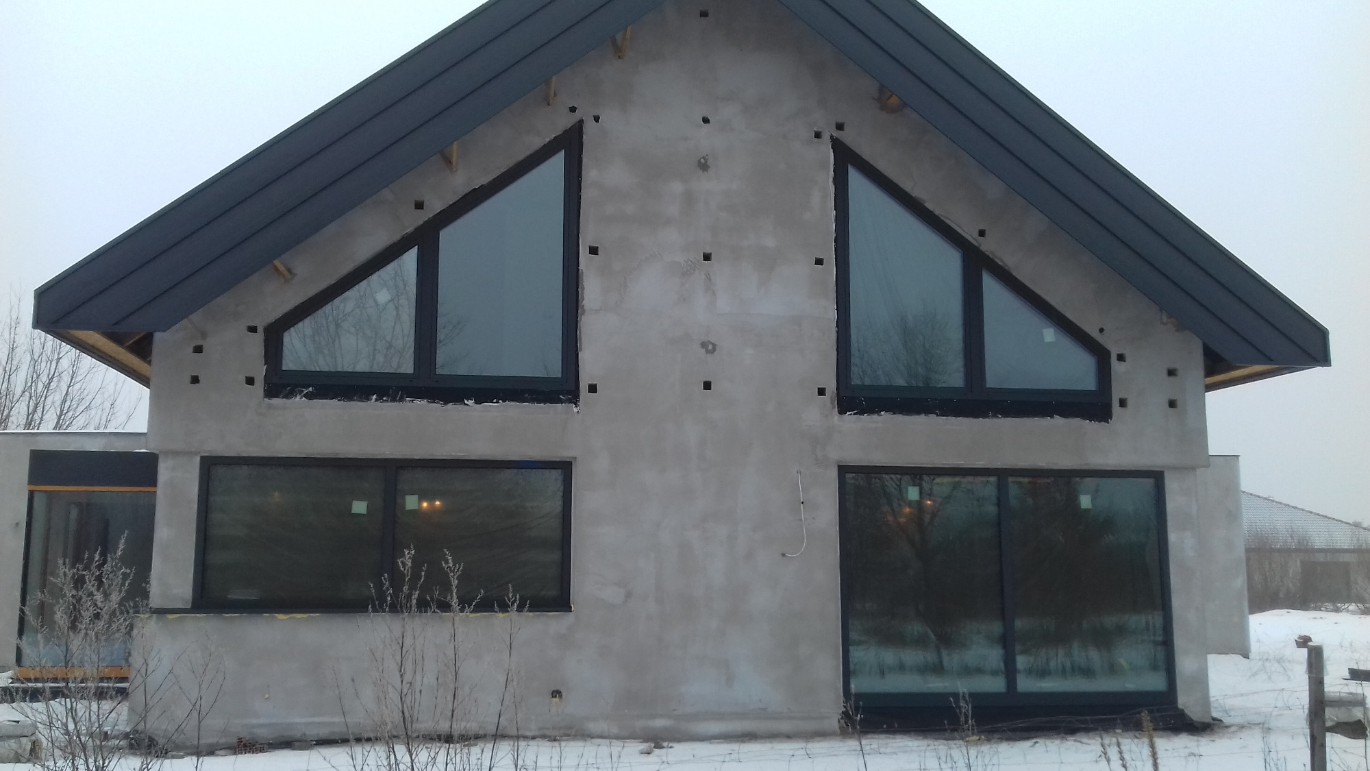 Surowy stan elewacji domu murowanego z dwoma trójkątnymi oknami na poddaszu i dwoma prostokątnymi na parterze, widoczne otwory wentylacyjne w ścianie, dach pokryty blachą w kolorze antracytowym...