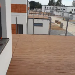 Nowoczesny taras z desek kompozytowych w kolorze brązowym, z metalową balustradą, widok na osiedle domów w zabudowie szeregowej.