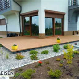 Nowoczesny, beżowy podest przed domem z brązowymi oknami, otoczony rabatami z kamyków i roślin, z żółtymi doniczkami z kwiatami.