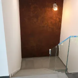 Wnętrze domu w trakcie wykańczania: schody z szarych płytek, szklana balustrada zabezpieczona taśmą malarską, ściana z dekoracyjną fakturą w odcieniach brązu, lampa wisząca zabezpieczona folią.