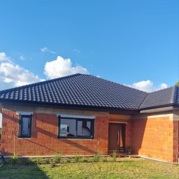 Firma Remontowo - Budowlana Sebastian Dziuda - Budowa domu jednorodzinnego z czerwonej cegły ceramicznej z czarną dachówką i oknami, widok z zewnątrz w słoneczny dzień.