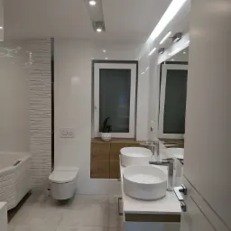 Nowoczesna łazienka z dwoma okrągłymi umywalkami nablatowymi, podwieszaną toaletą i wanną obudowaną płytkami z delikatnym wzorem, oświetlona ledowymi lampami sufitowymi i listwą świetlną nad lustrem.