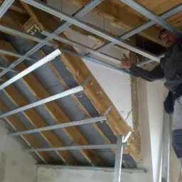 Montaż metalowego rusztu sufitowego na poddaszu z widoczną izolacją, pracownik na drabinie pokazuje kciuk w górę.