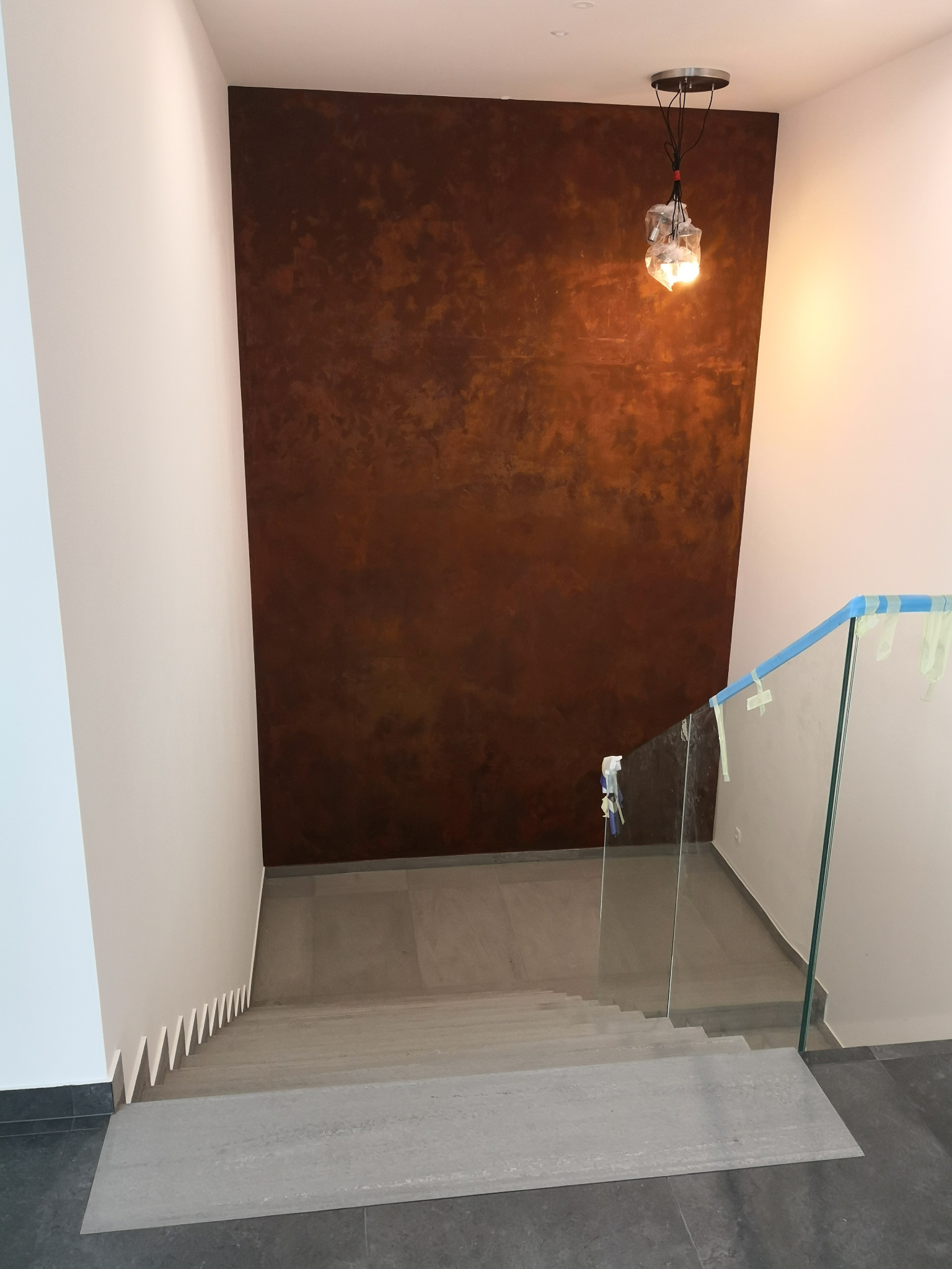 Wnętrze domu w trakcie wykańczania: schody z szarych płytek, szklana balustrada zabezpieczona taśmą malarską, ściana z dekoracyjną fakturą w odcieniach brązu, lampa wisząca zabezpieczona folią.