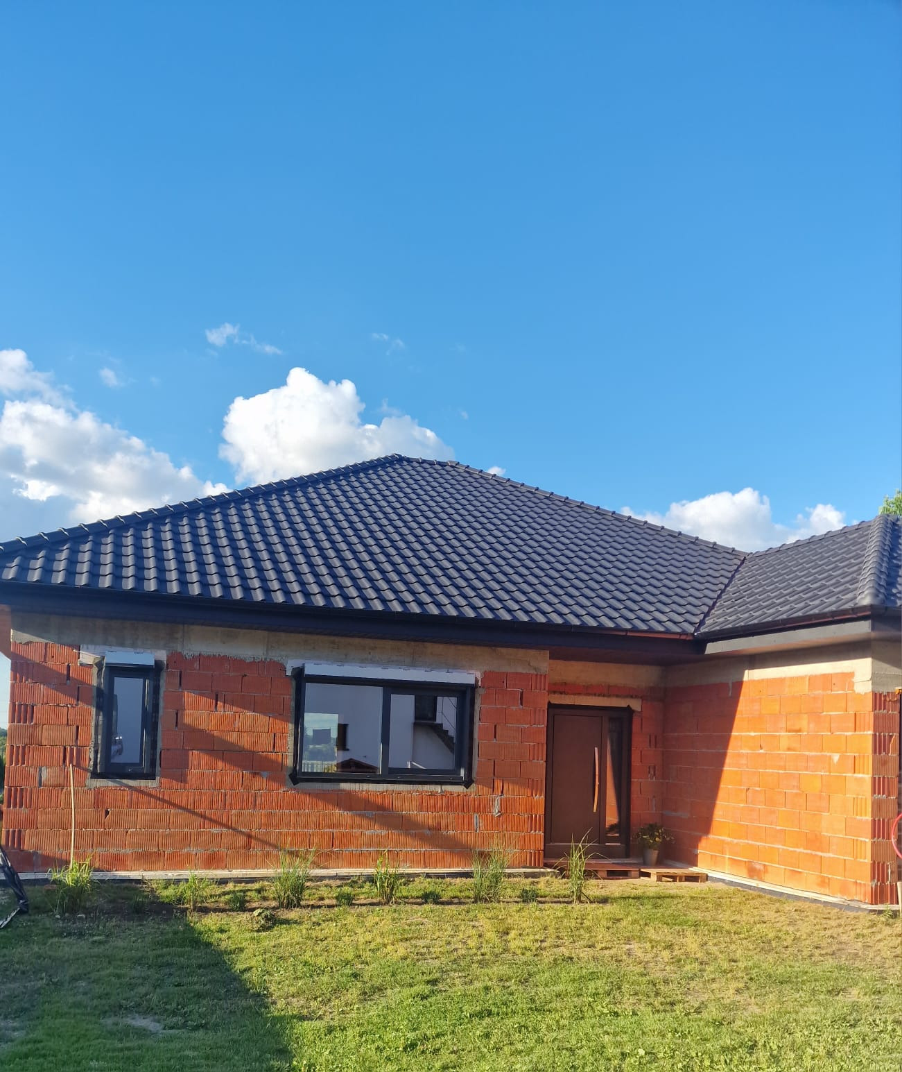 Budowa domu jednorodzinnego z czerwonej cegły ceramicznej z czarną dachówką i oknami, widok z zewnątrz w słoneczny dzień.