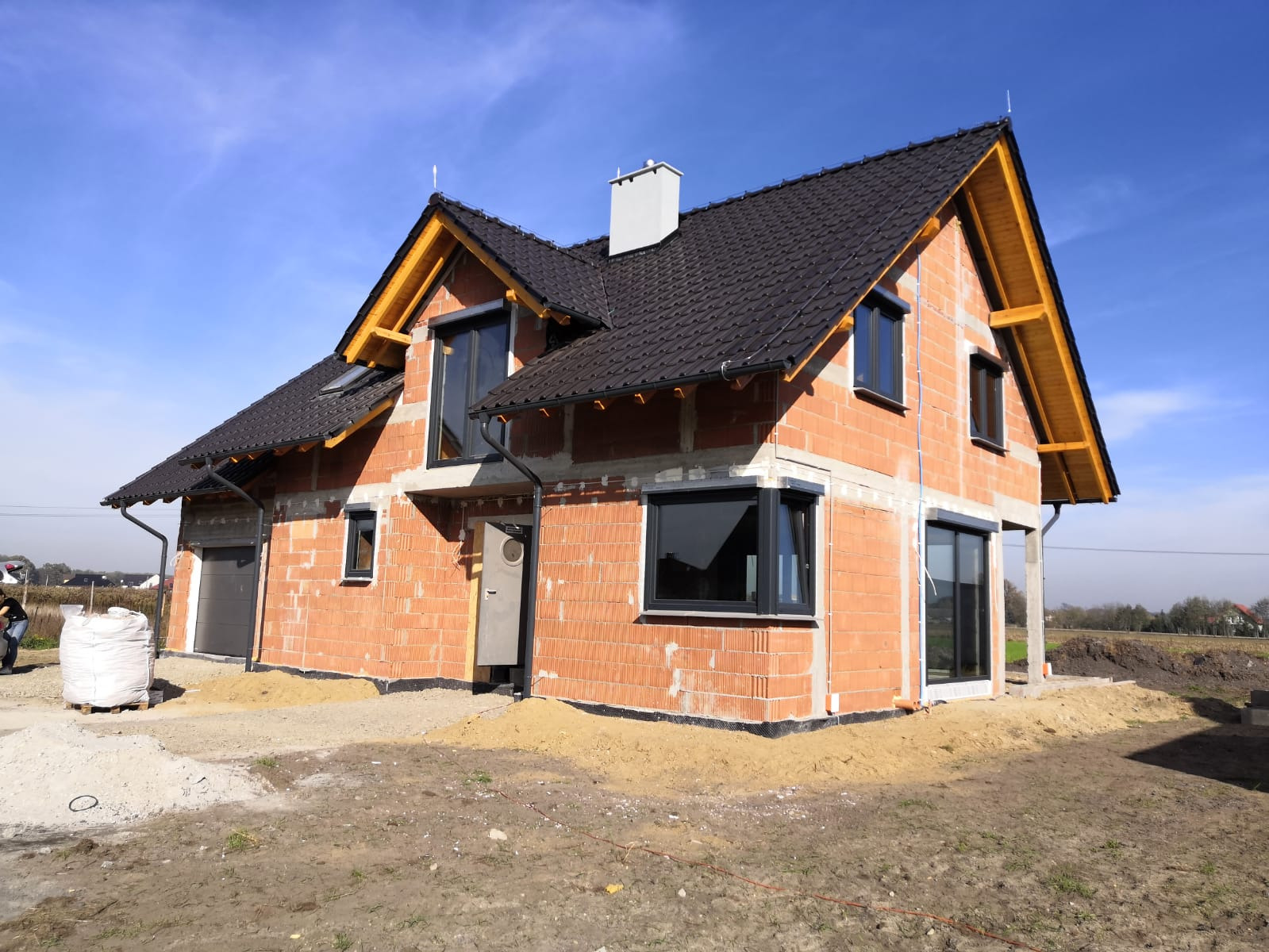 Dwukondygnacyjny dom w trakcie budowy, widoczne ściany z czerwonej cegły, czarne okna, dachówka i szary komin, teren wokół domu częściowo wyrównany.