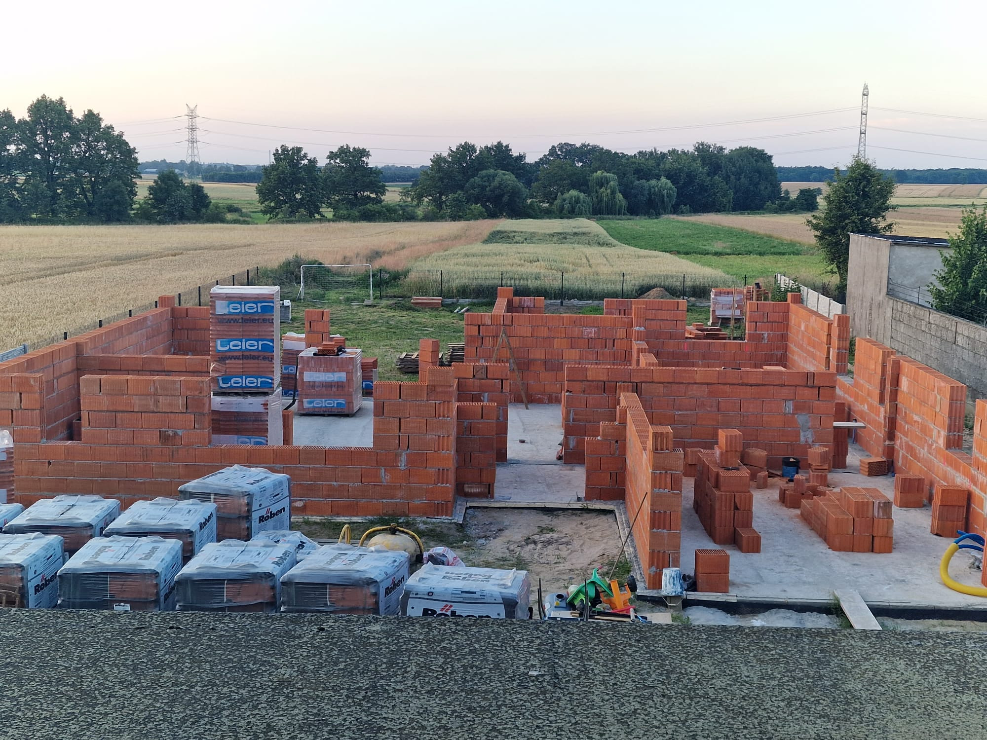 Wznoszenie ścian parteru domu z czerwonej cegły, widoczne palety z cegłami marki Roben i Leier, w tle pola i słupy wysokiego napięcia.