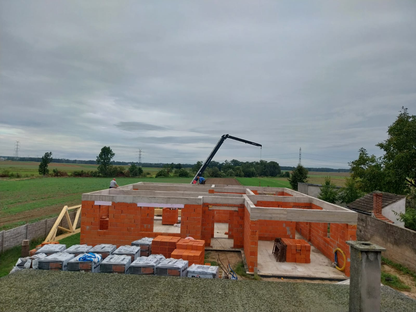 Budowa domu murowanego w trakcie: ściany z czerwonej cegły, betonowe nadproża, palety z cegłami, żuraw budowlany w tle, widok z góry na plac budowy.