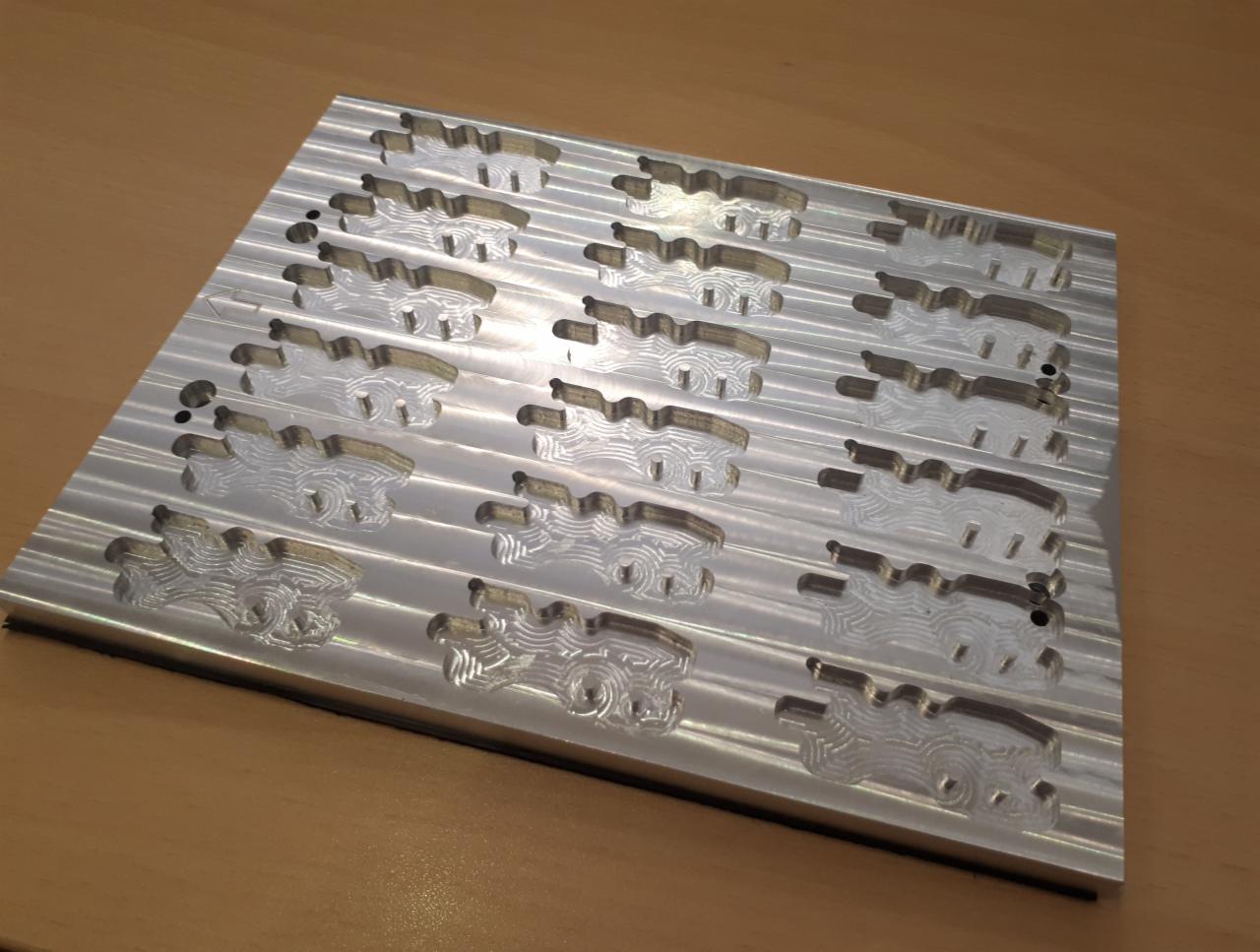 Aluminiowa płyta z precyzyjnie wyfrezowanymi, powtarzalnymi wzorami w kształcie zbliżonym do aut, wykonana techniką CNC, leżąca na drewnianym blacie.