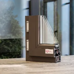 Przekrój brązowej ramy okiennej z trzema szybami, widoczne okucia i uszczelki, na drewnianym blacie, w tle zieleń ogrodu za oknem.