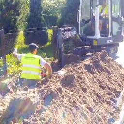 Pracownik w kamizelce odblaskowej Technopol, kopie rów obok koparki Terex TC16, na tle zieleni i ogrodzenia.