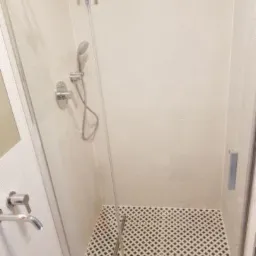 Nowoczesna kabina prysznicowa z chromowaną armaturą i kratkowaną posadzką w jasnej łazience z białymi płytkami.