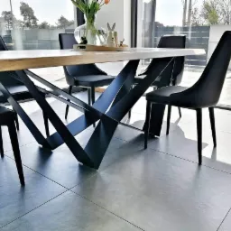 Stół z blatem z jasnego drewna i geometryczną, czarną podstawą z metalu, otoczony czarnymi krzesłami na tle jasnego wnętrza z dużym oknem.