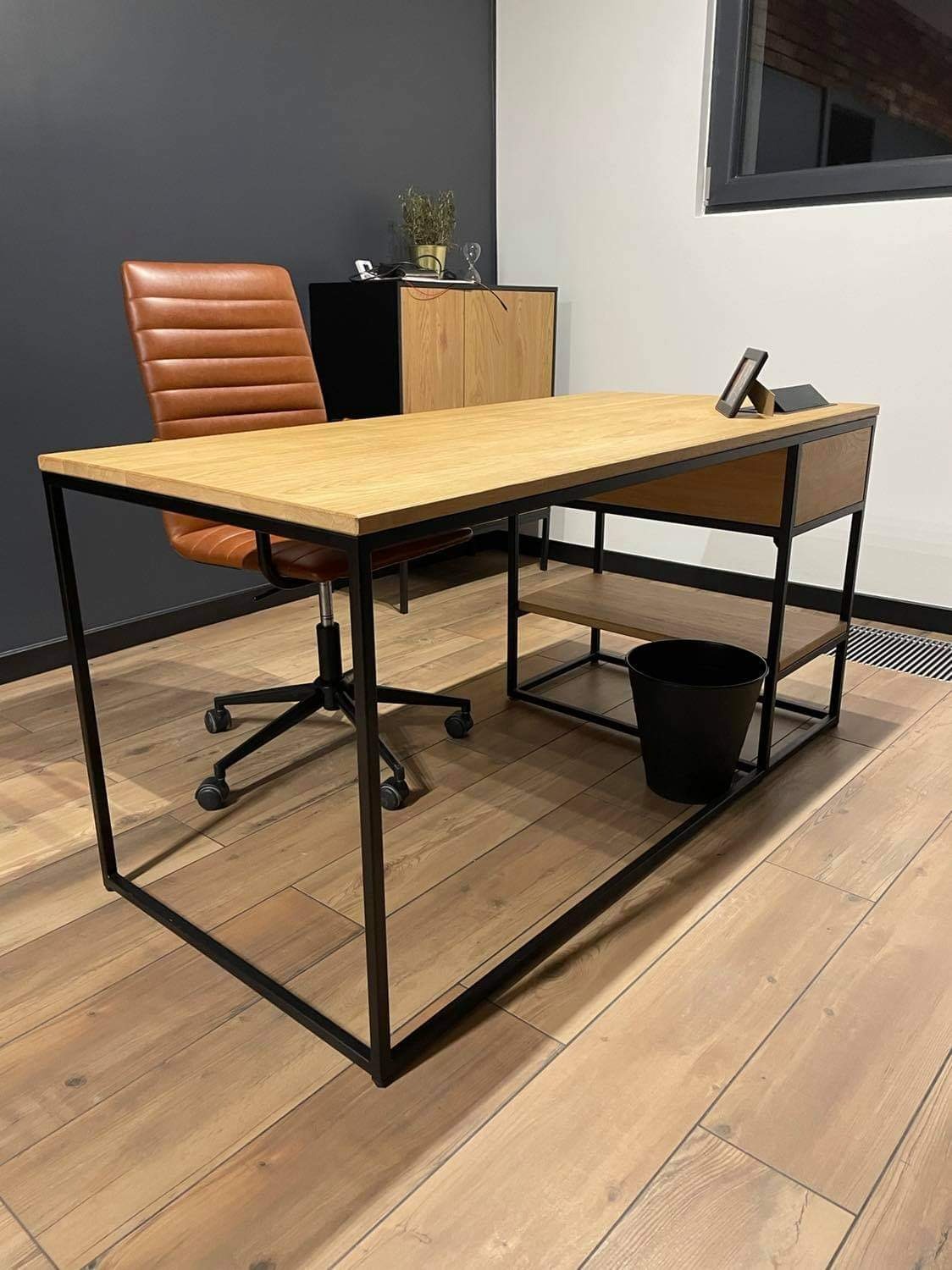 Biurko z jasnego drewna z czarną metalową ramą i półkami, brązowe krzesło biurowe na kółkach, czarny kosz na śmieci, drewniana podłoga, minimalistyczny wystrój biura.