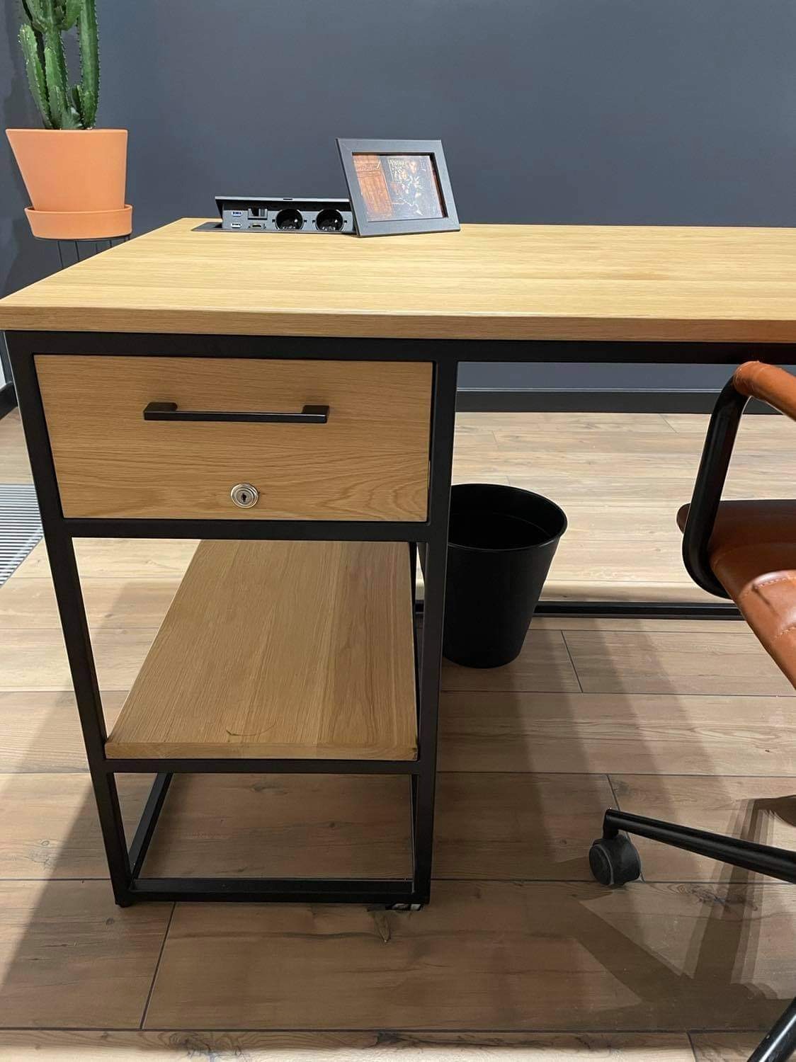 Biurko w stylu loft z drewnianym blatem i czarną metalową ramą, z szufladą zamykaną na klucz, półką i wbudowanym gniazdkiem elektrycznym, obok stoi skórzane krzesło na kółkach.