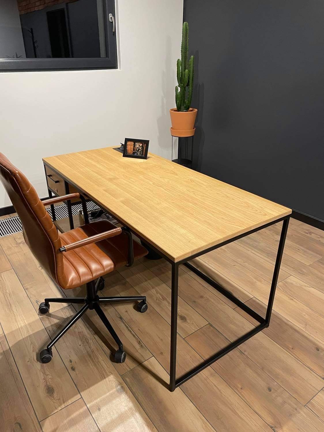 Nowoczesne biurko z jasnym drewnianym blatem i czarną metalową ramą, skórzane krzesło biurowe w odcieniach brązu, kaktus w doniczce na stojaku oraz ramka ze zdjęciem na blacie, na tle ściany...
