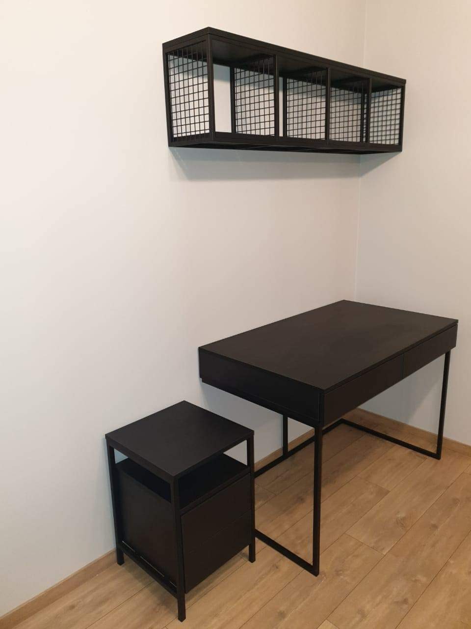 Czarny metalowy regał wiszący z siatkowymi ściankami nad biurkiem i szafką nocną w minimalistycznym stylu.