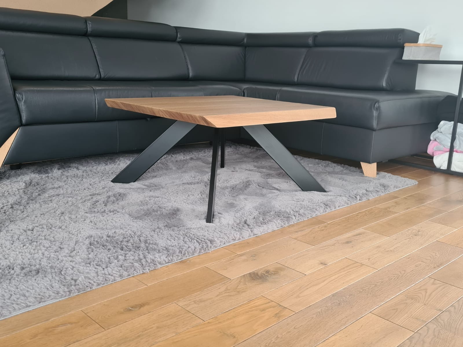 Kwadratowy stolik kawowy z drewnianym blatem i czarnymi, metalowymi nogami w kształcie litery X, ustawiony na puszystym, szarym dywanie, obok czarnej, skórzanej sofy narożnej.