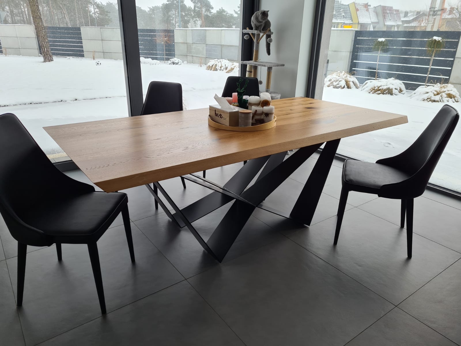 Drewniany stół jadalny z czarnymi, metalowymi nogami w nowoczesnym wnętrzu z widokiem na zimowy krajobraz za oknem. Na stole dekoracje w drewnianej tacy. Szary kot na drapaku w tle.