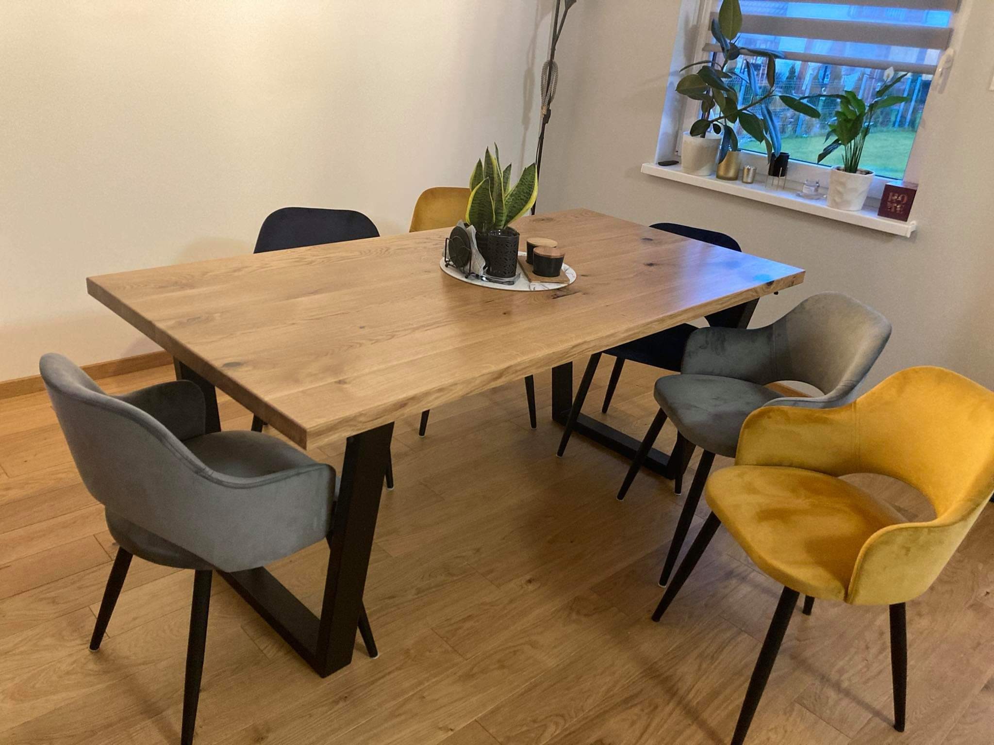 Stół z blatem z litego drewna na metalowych nogach w kształcie litery V, otoczony krzesłami w odcieniach szarości i musztardowej żółci, na blacie dekoracja z rośliną w doniczce.