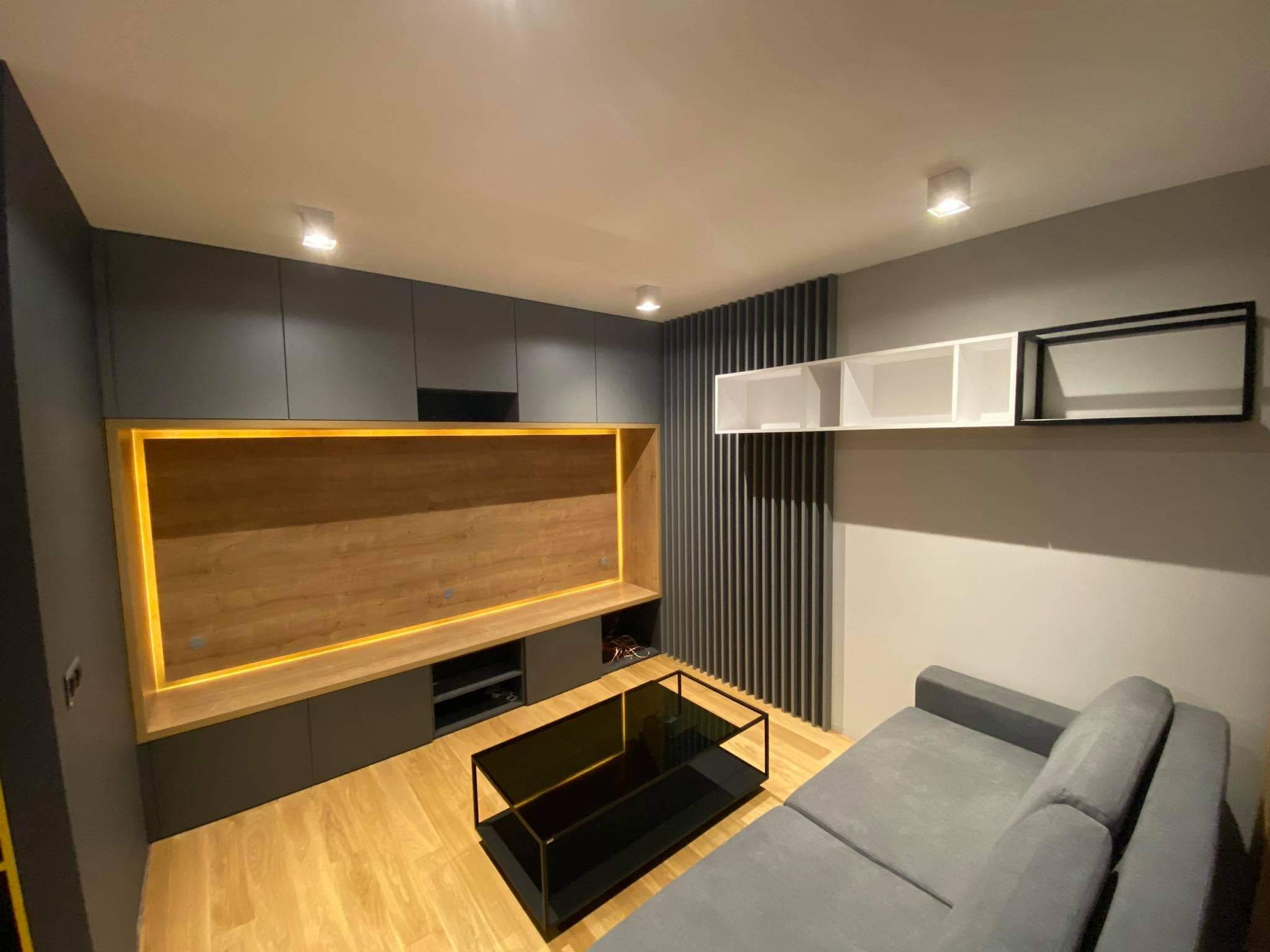 Nowoczesny salon z szarą zabudową meblową pod telewizor z drewnianym panelem podświetlonym LED, czarnym stolikiem kawowym i szarą sofą.