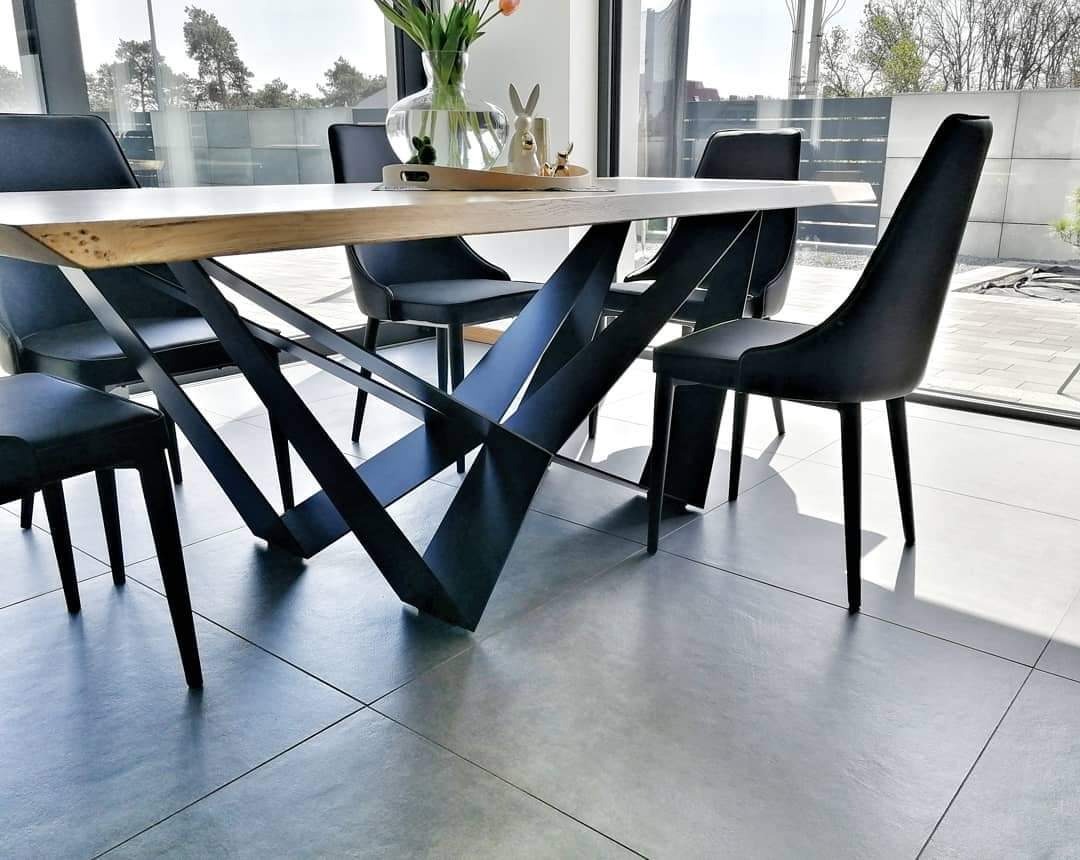 Stół z blatem z jasnego drewna i geometryczną, czarną podstawą z metalu, otoczony czarnymi krzesłami na tle jasnego wnętrza z dużym oknem.