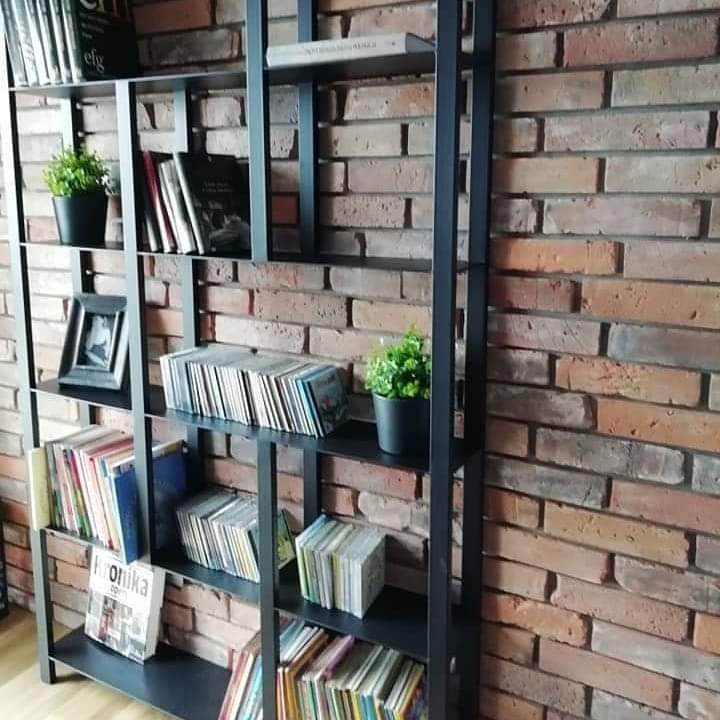 Czarny, metalowy regał na książki z półkami, ustawiony na tle ściany z cegły, wypełniony książkami, płytami CD, ramką ze zdjęciem i doniczkami z zielonymi roślinami.